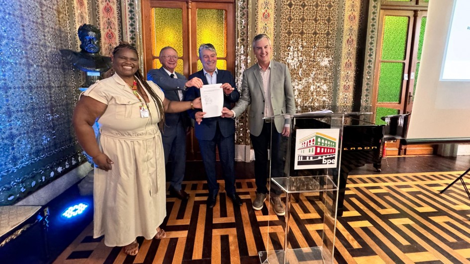 Esta &eacute; uma descri&ccedil;&atilde;o da imagem para fins de acessibilidade:

Descri&ccedil;&atilde;o da Imagem
Uma fotografia colorida em plano m&eacute;dio captura quatro pessoas (uma mulher e tr&ecirc;s homens) posando para uma foto oficial em um sal&atilde;o hist&oacute;rico da Biblioteca P&uacute;blica do Estado (BPE), em Porto Alegre.

A&ccedil;&atilde;o Principal: O grupo est&aacute; alinhado horizontalmente no centro da sala, segurando juntos um documento oficial branco em sinal de celebra&ccedil;&atilde;o por uma assinatura de conv&ecirc;nio ou repasse de recursos.

Os Participantes:

&Agrave; esquerda, uma mulher negra com longas tran&ccedil;as, vestindo um vestido bege de mangas curtas e sand&aacute;lias, sorri para a c&acirc;mera.

Ao centro e &agrave; direita, tr&ecirc;s homens em trajes casuais-finos (palet&oacute;s e camisas sociais em tons de azul e cinza) seguram as bordas do documento.

O Ambiente:

O sal&atilde;o &eacute; ricamente decorado em estilo cl&aacute;ssico. As paredes apresentam mosaicos ou pinturas intrincadas com padr&otilde;es geom&eacute;tricos e florais em tons de verde, vermelho e dourado.

Ao fundo, h&aacute; duas grandes portas ou janelas em arco com vitrais em tons de amarelo e verde.

&Agrave; esquerda, sobre um pedestal, v&ecirc;-se um busto de bronze.

&Agrave; direita, h&aacute; um piano de cauda preto e, &agrave; frente do grupo, um p&uacute;lpito de acr&iacute;lico transparente com o logotipo colorido da "BPE".

O ch&atilde;o possui um piso de madeira (parquet) com um padr&atilde;o geom&eacute;trico complexo que lembra um labirinto.

Ilumina&ccedil;&atilde;o: A luz &eacute; quente e interna, com um foco de luz azul vis&iacute;vel no ch&atilde;o, no canto inferior esquerdo.