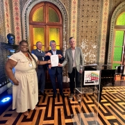 Esta &eacute; uma descri&ccedil;&atilde;o da imagem para fins de acessibilidade:

Descri&ccedil;&atilde;o da Imagem
Uma fotografia colorida em plano m&eacute;dio captura quatro pessoas (uma mulher e tr&ecirc;s homens) posando para uma foto oficial em um sal&atilde;o hist&oacute;rico da Biblioteca P&uacute;blica do Estado (BPE), em Porto Alegre.

A&ccedil;&atilde;o Principal: O grupo est&aacute; alinhado horizontalmente no centro da sala, segurando juntos um documento oficial branco em sinal de celebra&ccedil;&atilde;o por uma assinatura de conv&ecirc;nio ou repasse de recursos.

Os Participantes:

&Agrave; esquerda, uma mulher negra com longas tran&ccedil;as, vestindo um vestido bege de mangas curtas e sand&aacute;lias, sorri para a c&acirc;mera.

Ao centro e &agrave; direita, tr&ecirc;s homens em trajes casuais-finos (palet&oacute;s e camisas sociais em tons de azul e cinza) seguram as bordas do documento.

O Ambiente:

O sal&atilde;o &eacute; ricamente decorado em estilo cl&aacute;ssico. As paredes apresentam mosaicos ou pinturas intrincadas com padr&otilde;es geom&eacute;tricos e florais em tons de verde, vermelho e dourado.

Ao fundo, h&aacute; duas grandes portas ou janelas em arco com vitrais em tons de amarelo e verde.

&Agrave; esquerda, sobre um pedestal, v&ecirc;-se um busto de bronze.

&Agrave; direita, h&aacute; um piano de cauda preto e, &agrave; frente do grupo, um p&uacute;lpito de acr&iacute;lico transparente com o logotipo colorido da "BPE".

O ch&atilde;o possui um piso de madeira (parquet) com um padr&atilde;o geom&eacute;trico complexo que lembra um labirinto.

Ilumina&ccedil;&atilde;o: A luz &eacute; quente e interna, com um foco de luz azul vis&iacute;vel no ch&atilde;o, no canto inferior esquerdo.