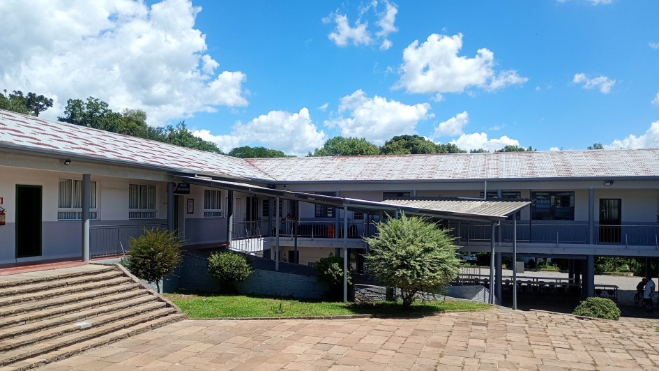 P&aacute;tio externo de uma escola com piso de blocos de concreto avermelhados. &Agrave; esquerda, escadaria ampla leva a um edif&iacute;cio t&eacute;rreo branco. Ao centro e &agrave; direita, pr&eacute;dio de dois andares com paredes brancas e detalhes azuis, corredores com grades met&aacute;licas e telhado cinza. Pequeno jardim com arbustos e &aacute;rvores ornamentais ao redor. Ao fundo, vegeta&ccedil;&atilde;o densa e c&eacute;u azul com nuvens brancas volumosas. Duas pessoas caminham no canto inferior direito.