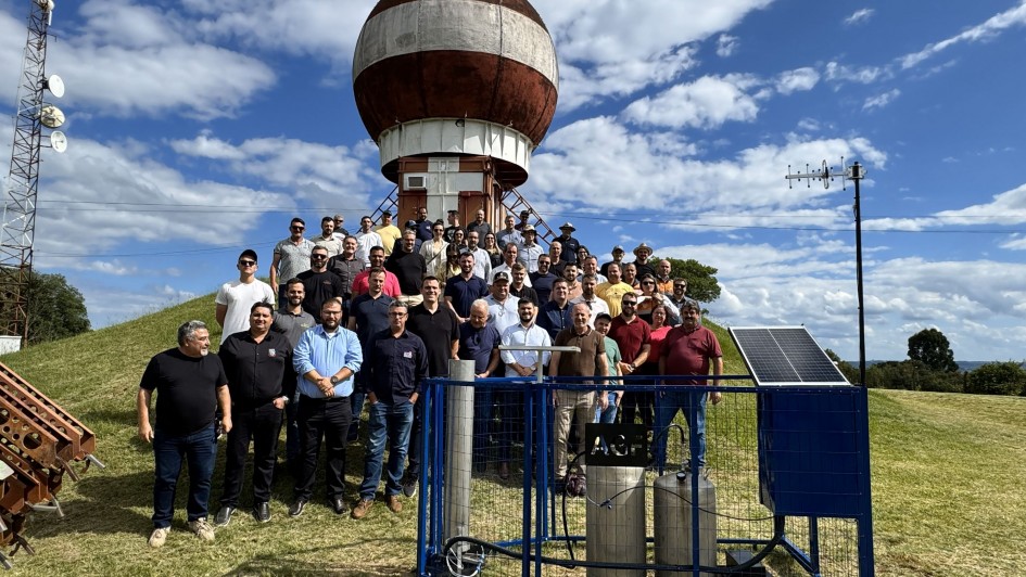 Foto posada ao ar livre de um grupo de mais de 50 pessoas de p&eacute;, em ambiente de campo aberto. Ao fundo h&aacute; uma estrutura redonda.