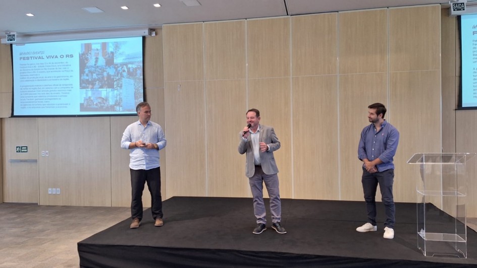 Foto de tr&ecirc;s homens, em trajes formais, de p&eacute; num palco baixo, localizado em ambiente fechado. Ao centro, Santini aparece falando no microfone.