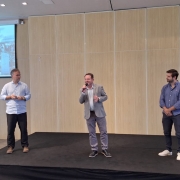 Foto de tr&ecirc;s homens, em trajes formais, de p&eacute; num palco baixo, localizado em ambiente fechado. Ao centro, Santini aparece falando no microfone.