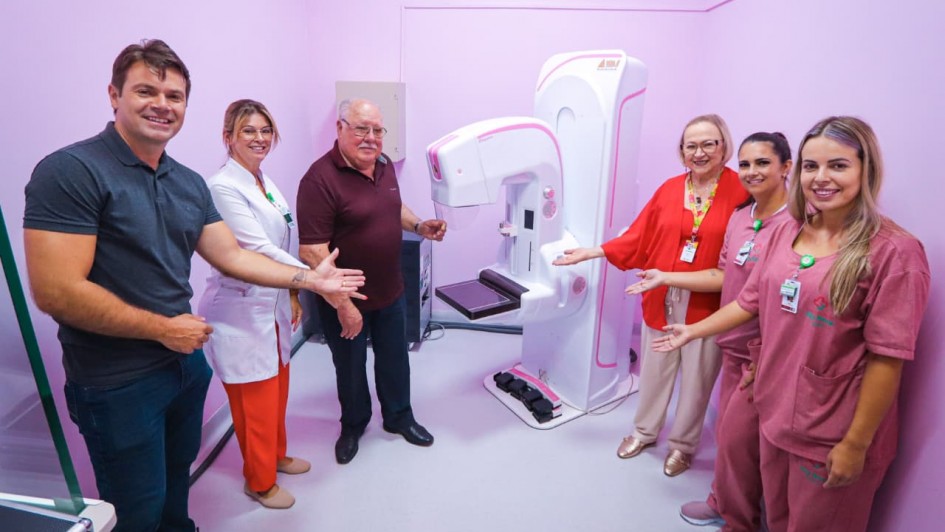 Descri&ccedil;&atilde;o da Imagem
Uma fotografia colorida em plano m&eacute;dio e &acirc;ngulo aberto captura a entrega de um novo equipamento m&eacute;dico em uma sala de exames no munic&iacute;pio de Tapes.

A&ccedil;&atilde;o Principal: Um grupo de seis pessoas (quatro mulheres e dois homens) est&aacute; posicionado ao redor de um mam&oacute;grafo moderno, fazendo gestos com as m&atilde;os abertas em dire&ccedil;&atilde;o ao aparelho, como em uma apresenta&ccedil;&atilde;o oficial. Todos sorriem para a c&acirc;mera.

O Equipamento: No centro da sala, destaca-se um mam&oacute;grafo de &uacute;ltima gera&ccedil;&atilde;o, predominantemente branco com detalhes em rosa. O aparelho possui uma base robusta e bra&ccedil;os mec&acirc;nicos ajust&aacute;veis para a realiza&ccedil;&atilde;o dos exames.

Os Participantes:

&Agrave; esquerda: Um homem de polo cinza escuro, uma mulher de jaleco branco sobre cal&ccedil;a laranja e um homem de polo bord&ocirc;.

&Agrave; direita: Uma mulher de blusa vermelha vibrante e cal&ccedil;a bege, seguida por duas mulheres usando uniformes privativos (scrubs) de cor rosa.

O Ambiente: A sala &eacute; pequena e possui todas as paredes pintadas em um tom de rosa claro, em alus&atilde;o &agrave; sa&uacute;de da mulher. No alto da parede ao fundo, v&ecirc;-se um aparelho de ar-condicionado branco. O piso &eacute; de um tom cinza claro neutro.
