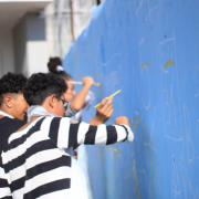 Esta &eacute; uma descri&ccedil;&atilde;o da imagem para fins de acessibilidade:

Descri&ccedil;&atilde;o da Imagem
Uma fotografia colorida em plano m&eacute;dio e &acirc;ngulo lateral captura tr&ecirc;s jovens participando de uma atividade de pintura art&iacute;stica em um muro escolar, parte do projeto Artista na Escola.

A&ccedil;&atilde;o Principal: Os jovens est&atilde;o alinhados ao longo de um muro azul vibrante, utilizando pinc&eacute;is finos de cabo amarelo para preencher ou contornar desenhos e n&uacute;meros previamente esbo&ccedil;ados em branco.

Os Jovens:

Em primeiro plano, um jovem de cabelos escuros e cacheados veste uma camiseta de mangas longas com listras horizontais pretas e brancas. Ele est&aacute; focado na pintura &agrave; sua frente.

Atr&aacute;s dele, um segundo jovem de perfil tamb&eacute;m trabalha no muro.

Ao fundo, uma terceira estudante com o cabelo preso em um coque alto &eacute; vista segurando um pincel.

O Muro: A superf&iacute;cie &eacute; de um tom azul claro intenso. Sobre ela, h&aacute; marca&ccedil;&otilde;es em giz ou tinta branca que lembram um calend&aacute;rio ou grade num&eacute;rica, com n&uacute;meros como "5", "24/31", "28" e "29" vis&iacute;veis.

O Ambiente:

A cena ocorre ao ar livre sob luz natural clara.

O fundo est&aacute; levemente desfocado, mostrando parte da estrutura cinza da escola e uma porta ou port&atilde;o de metal branco com grades.

A profundidade de campo &eacute; reduzida, mantendo o foco principal no jovem com a camiseta listrada e em sua a&ccedil;&atilde;o de pintar.