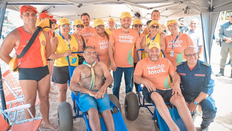Foto mostra o vice-governador, Gabriel Souza, junto com outros integrantes da Opera&ccedil;&atilde;o Ver&atilde;o Total, na praia do Cassino, que vestem camisetas laranjas do programa. H&aacute; tamb&eacute;m um salva-vidas e um bombeiro. Todos est&atilde;o perfilados para foto embaixo de uma barraca na areia, atr&aacute;s de dois cadeirantes que est&atilde;o em cadeiras de rodas adaptadas para entrar no mar. 