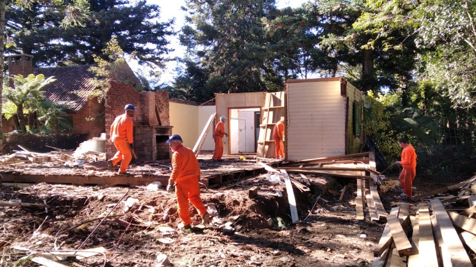 Descri&ccedil;&atilde;o da Imagem
Uma fotografia colorida em plano aberto captura cinco trabalhadores em uma for&ccedil;a-tarefa de desmonte de uma estrutura de madeira em uma &aacute;rea densamente arborizada.

A&ccedil;&atilde;o Principal: Os trabalhadores, vestindo uniformes de cor laranja vibrante, est&atilde;o espalhados pelo canteiro de obras. Eles trabalham na demoli&ccedil;&atilde;o e remo&ccedil;&atilde;o de materiais de uma casa de madeira parcialmente destru&iacute;da. Dois trabalhadores caminham carregando ferramentas ou pe&ccedil;as, enquanto outros tr&ecirc;s est&atilde;o posicionados pr&oacute;ximos &agrave;s paredes restantes da estrutura.

Os Trabalhadores: Todos vestem macac&otilde;es de pe&ccedil;a &uacute;nica na cor laranja, capacetes de prote&ccedil;&atilde;o azuis e luvas de trabalho. Suas posturas indicam esfor&ccedil;o f&iacute;sico e coordena&ccedil;&atilde;o na tarefa.

A Estrutura:

Trata-se de uma constru&ccedil;&atilde;o de madeira com paredes de t&aacute;buas horizontais em tom bege claro.

Grande parte do telhado e das paredes j&aacute; foi removida, restando uma base de madeira e uma chamin&eacute; de tijolos aparentes com uma lareira na base, que permanece em p&eacute; em meio aos escombros.

O ch&atilde;o est&aacute; coberto por terra batida, folhagens e muitas ripas e t&aacute;buas de madeira espalhadas.

O Ambiente: * A cena ocorre em um local cercado por &aacute;rvores altas e frondosas, com destaque para pinheiros que filtram a luz do sol.

A ilumina&ccedil;&atilde;o &eacute; natural e intensa, criando contrastes marcantes de luz e sombra no terreno e sobre os uniformes dos trabalhadores.

Ao fundo, &agrave; esquerda, &eacute; poss&iacute;vel ver o telhado de outra constru&ccedil;&atilde;o de estilo r&uacute;stico que permanece intacta.