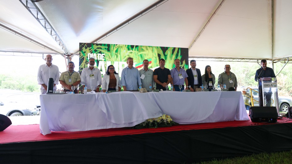 Descri&ccedil;&atilde;o Geral: A imagem mostra um grupo de autoridades e representantes reunidos em um palco sob uma grande tenda branca durante um evento oficial ao ar livre.

Detalhes da Imagem:

O Palco: O palco possui um piso coberto por um tapete vermelho e uma saia lateral de tecido preto. Na parte frontal, h&aacute; uma longa mesa forrada com uma toalha branca que chega at&eacute; o ch&atilde;o. Sobre a mesa, veem-se garrafas de &aacute;gua, copos e pequenas placas de identifica&ccedil;&atilde;o.

As Pessoas: Onze pessoas (dez homens e uma mulher) est&atilde;o posicionadas em p&eacute; atr&aacute;s da mesa, voltadas para a c&acirc;mera. Elas vestem roupas casuais e formais, como camisas sociais e jaquetas leves. &Agrave; direita do grupo, um homem est&aacute; posicionado em um p&uacute;lpito de acr&iacute;lico transparente com o logotipo do IRGA (Instituto Rio Grandense do Arroz), falando ao microfone.

O Painel: Ao fundo, atr&aacute;s das pessoas, h&aacute; um grande tel&atilde;o digital exibindo a identidade visual do evento. &Eacute; poss&iacute;vel ler as palavras "DIA DE CAMPO" em letras grandes e brancas, sobrepostas a uma imagem estilizada de planta&ccedil;&otilde;es verdes.

O Ambiente: O evento ocorre sob uma estrutura de tenda met&aacute;lica com lona branca. Ao fundo, nas laterais, vislumbra-se a vegeta&ccedil;&atilde;o e parte de ve&iacute;culos estacionados, indicando um ambiente rural ou de campo. O ch&atilde;o &agrave; frente do palco &eacute; de grama verde.