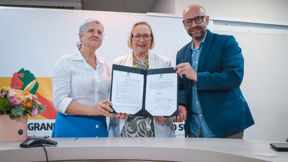 Esta &eacute; uma descri&ccedil;&atilde;o da imagem para fins de acessibilidade:

Descri&ccedil;&atilde;o Geral: Tr&ecirc;s pessoas em um ambiente formal interno posam sorridentes para a foto, segurando juntas uma pasta aberta com documentos oficiais assinados.

Detalhes da Imagem:

As Pessoas:

&Agrave; esquerda, uma mulher de cabelos grisalhos presos, usando &oacute;culos e vestindo uma blusa branca de bot&otilde;es com uma saia azul.

No centro, uma mulher de cabelos loiros curtos, usando &oacute;culos, uma blusa com estampa verde e branca e um blazer branco por cima.

&Agrave; direita, um homem calvo, de barba e &oacute;culos, vestindo um blazer azul sobre uma camisa azul clara.

O Documento: Os tr&ecirc;s seguram uma pasta preta aberta que exibe duas p&aacute;ginas de uma portaria ou documento oficial, com textos e assinaturas vis&iacute;veis.

O Primeiro Plano: &Agrave; frente deles, h&aacute; uma mesa curva de cor clara. Sobre a mesa, no canto esquerdo, v&ecirc;-se um pequeno arranjo de flores coloridas em um vaso rosa e um microfone de mesa preto.

O Plano de Fundo: O fundo &eacute; composto por uma parede branca e partes de banners promocionais. No lado esquerdo, aparece um logotipo colorido e o in&iacute;cio da palavra "GRAN".
