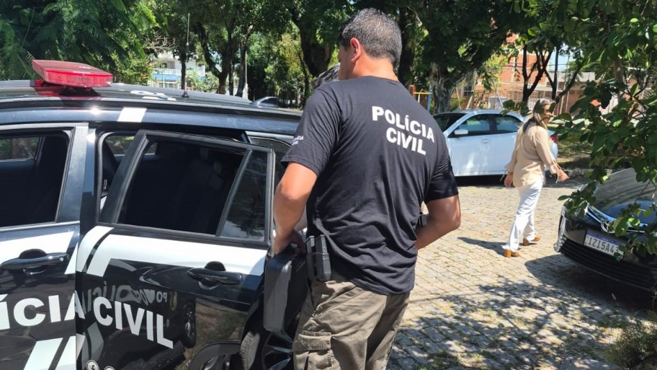 A imagem mostra um policial civil perto de uma viatura. Ele veste uma camiseta de uniforme de cor preta e est&aacute; ao ar livre, em uma rua com outros carros e &aacute;rvores.