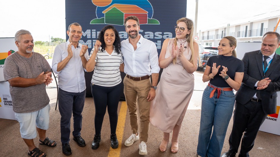 Esta &eacute; uma descri&ccedil;&atilde;o da imagem para fins de acessibilidade:

Descri&ccedil;&atilde;o Geral: O governador Eduardo Leite e outras autoridades posam ao lado de uma benefici&aacute;ria que segura as chaves de sua nova casa durante um evento oficial de entrega de moradias em Eldorado do Sul.

Detalhes da Imagem:

As Pessoas: No centro da foto, o governador Eduardo Leite (homem jovem, de barba, vestindo camisa branca social e cal&ccedil;a bege) sorri com o bra&ccedil;o sobre o ombro de uma mulher. Esta mulher, a benefici&aacute;ria, veste uma camiseta listrada em preto e branco e sorri enquanto exibe um molho de chaves para a c&acirc;mera.

O Grupo: Ao redor deles, outras cinco pessoas (tr&ecirc;s homens e duas mulheres) aplaudem e sorriem. Os trajes variam entre o casual (camisetas e jeans) e o formal (um homem &agrave; direita veste palet&oacute; e gravata).

O Painel: Logo atr&aacute;s do grupo, h&aacute; um grande banner azul escuro com o logotipo de um programa habitacional, mostrando uma casa estilizada colorida sob um sol, com parte do texto "Minha Casa" vis&iacute;vel.

O Ambiente: O grupo est&aacute; sob uma estrutura de cobertura met&aacute;lica. Ao fundo, no lado direito, &eacute; poss&iacute;vel ver uma fileira de casas novas geminadas, pintadas de branco com detalhes em cinza, representando o conjunto habitacional entregue. O dia parece claro e ensolarado.