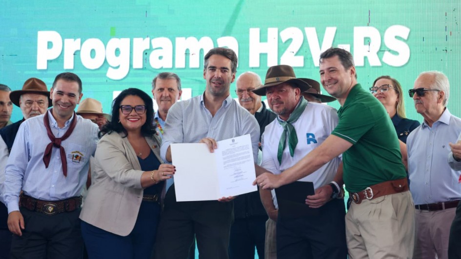 Um grupo de pessoas posa sorrindo para a foto enquanto segura um documento, em frente a um painel que diz &ldquo;Programa H2V RS&rdquo;.