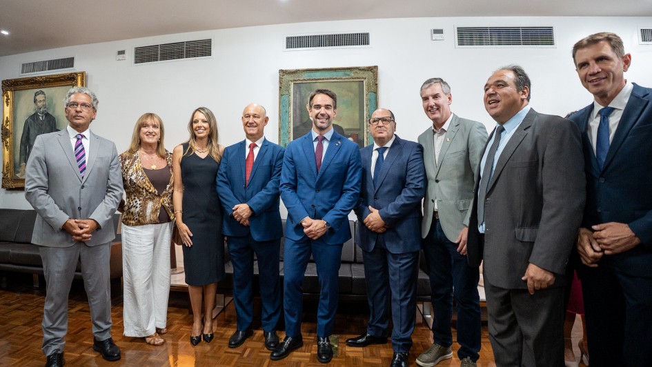 Esta &eacute; uma fotografia oficial de um evento pol&iacute;tico institucional, capturada em um ambiente interno com ilumina&ccedil;&atilde;o clara e formal.

Descri&ccedil;&atilde;o da Imagem
A imagem apresenta um grupo de dez pessoas (duas mulheres e oito homens) posando lado a lado para uma foto oficial. Eles est&atilde;o dispostos em uma linha horizontal, ocupando o centro do quadro, em uma sala com piso de taco de madeira e paredes brancas.

Detalhes dos Participantes
Centro: O Governador do Rio Grande do Sul, Eduardo Leite, destaca-se ao centro. Ele veste um terno azul vibrante, camisa social azul clara e uma gravata bord&ocirc;. Ele sorri para a c&acirc;mera com as m&atilde;os cruzadas &agrave; frente do corpo.

Vestimentas: * A maioria dos homens utiliza trajes formais (ternos em tons de cinza, azul-marinho e preto com gravatas variadas).

Uma das mulheres veste uma blusa com estampa de animal print (on&ccedil;a), cal&ccedil;a branca e sand&aacute;lias.

A outra mulher usa um vestido preto cl&aacute;ssico de comprimento m&eacute;dio e sapatos de salto alto.

Postura: Todos est&atilde;o de p&eacute;, com express&otilde;es cordiais e sorridentes, sugerindo um clima de celebra&ccedil;&atilde;o e formalidade.

O Cen&aacute;rio
Paredes: Ao fundo, h&aacute; duas pinturas emolduradas. &Agrave; esquerda, um retrato cl&aacute;ssico de um homem com barba; ao centro-fundo, uma obra de arte mais abstrata com tons esverdeados.

Mobili&aacute;rio: Um sof&aacute; de couro escuro est&aacute; posicionado logo atr&aacute;s do grupo. Na parte superior da parede, &eacute; poss&iacute;vel ver as grelhas do sistema de ar-condicionado central.

Piso: O ch&atilde;o &eacute; de madeira encerada, refletindo levemente a luz do ambiente e as silhuetas das pessoas.