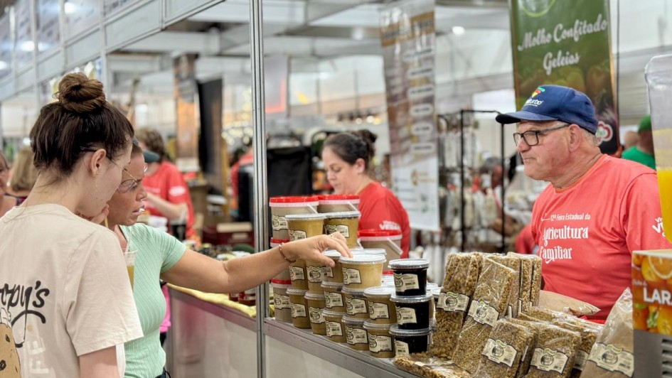 A imagem mostra uma banca de produtos de cana-de-a&ccedil;&uacute;car, como melado e rapadura. Duas mulheres observam e tocam os potes, enquanto duas pessoas est&atilde;o do outro lado da banca. 