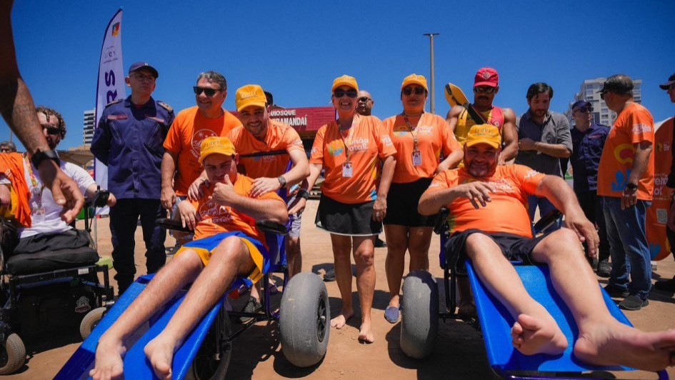 Esta &eacute; uma descri&ccedil;&atilde;o detalhada da imagem para fins de acessibilidade:

Descri&ccedil;&atilde;o Geral
A fotografia, tirada sob a luz forte do dia em uma praia, mostra um grupo de cerca de dez pessoas reunidas na areia para uma a&ccedil;&atilde;o de acessibilidade. O clima &eacute; ensolarado e positivo.

Elementos em Destaque
Primeiro Plano: Dois homens est&atilde;o sentados em cadeiras de rodas anf&iacute;bias de cor azul vibrante, equipadas com grandes rodas cinzas emborrachadas, pr&oacute;prias para deslocamento na areia. Ambos vestem camisetas laranjas. O homem &agrave; esquerda sorri e faz um sinal de "positivo" (polegar para cima) com a m&atilde;o.

Grupo de Apoio: Atr&aacute;s dos cadeirantes, v&aacute;rias pessoas est&atilde;o em p&eacute;. A maioria veste camisetas laranjas e bon&eacute;s amarelos (uniforme do projeto "Ver&atilde;o Total"). Entre eles, destacam-se:

Um homem &agrave; esquerda com uniforme azul-marinho do Corpo de Bombeiros.

Um salva-vidas ao fundo, usando uma regata vermelha e amarela, segurando um equipamento de resgate.

Homens e mulheres sorridentes, posicionados como equipe de apoio ou autoridades.

Cen&aacute;rio e Fundo
Ch&atilde;o: Areia clara e batida de praia.

Fundo: H&aacute; uma estrutura vermelha com a inscri&ccedil;&atilde;o "QUIOSQUE TRAMANDA&Iacute;". Ao fundo, o c&eacute;u &eacute; de um azul l&iacute;mpido, sem nuvens. &Agrave; esquerda, v&ecirc;-se uma bandeira vertical roxa e branca.

Ilumina&ccedil;&atilde;o: A luz solar &eacute; intensa, criando sombras curtas e real&ccedil;ando as cores vibrantes das roupas e dos equipamentos.