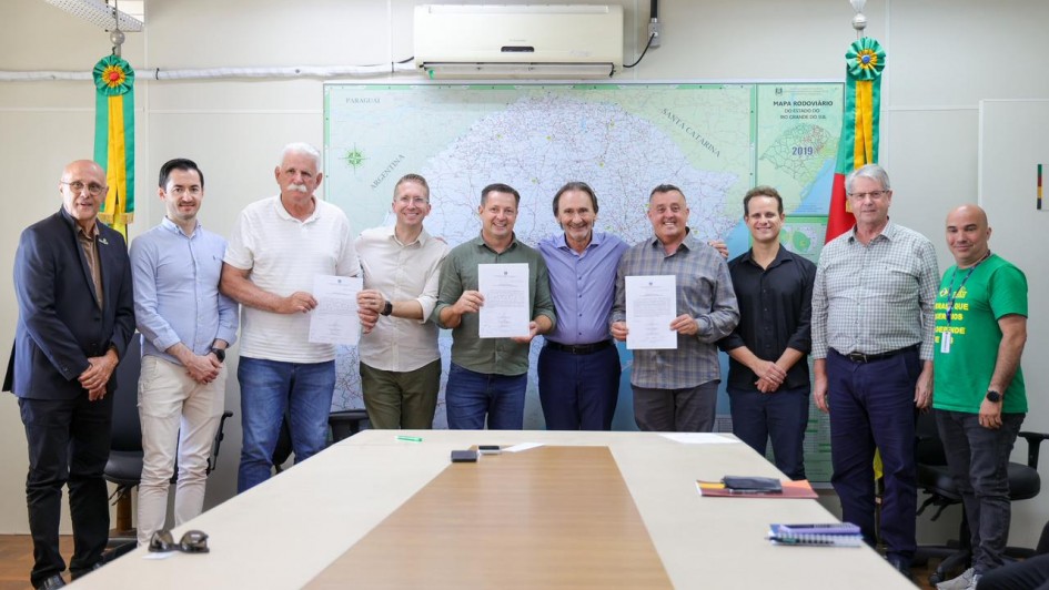 A imagem mostra um grupo de dez homens em p&eacute;, reunidos em frente a uma mesa de reuni&otilde;es. Tr&ecirc;s deles seguram documentos assinados. Ao fundo, h&aacute; um mapa do Rio Grande do Sul. 