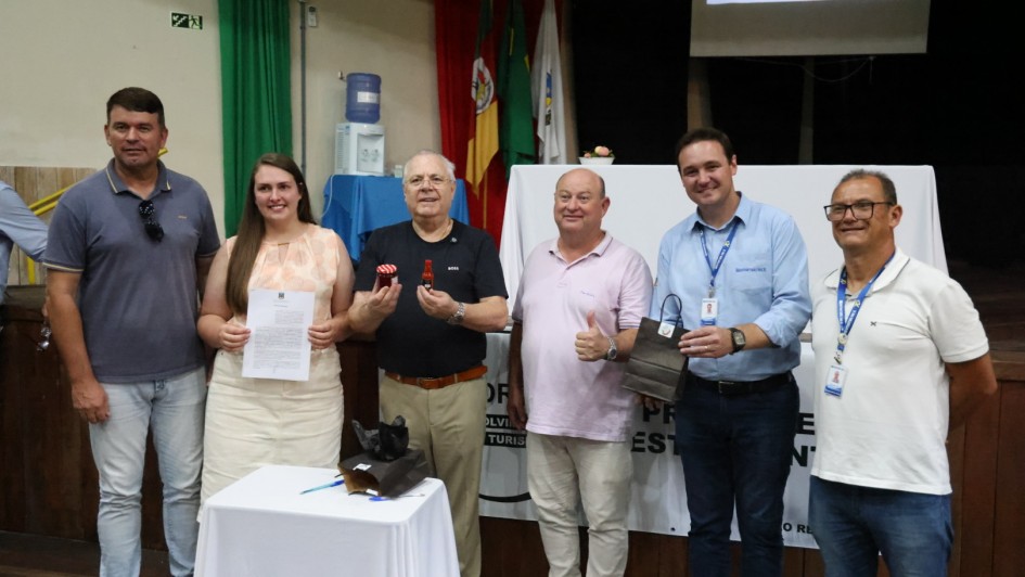 Um grupo de seis pessoas reunidas, em p&eacute;, posando para a foto em um ambiente fechado. H&aacute; uma mesa ao centro e uma pessoa segura um documento assinado. Ao fundo, h&aacute; a bandeira do RS. 