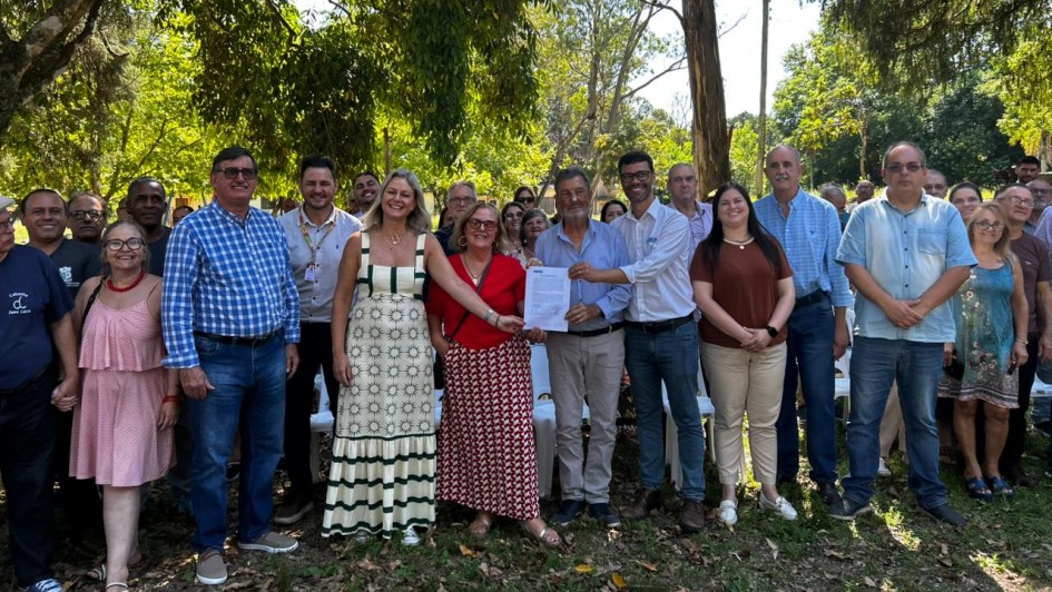 Um grupo de pessoas est&aacute; em p&eacute; ao ar livre posando para a foto. No centro, duas pessoas seguram um documento assinado. 