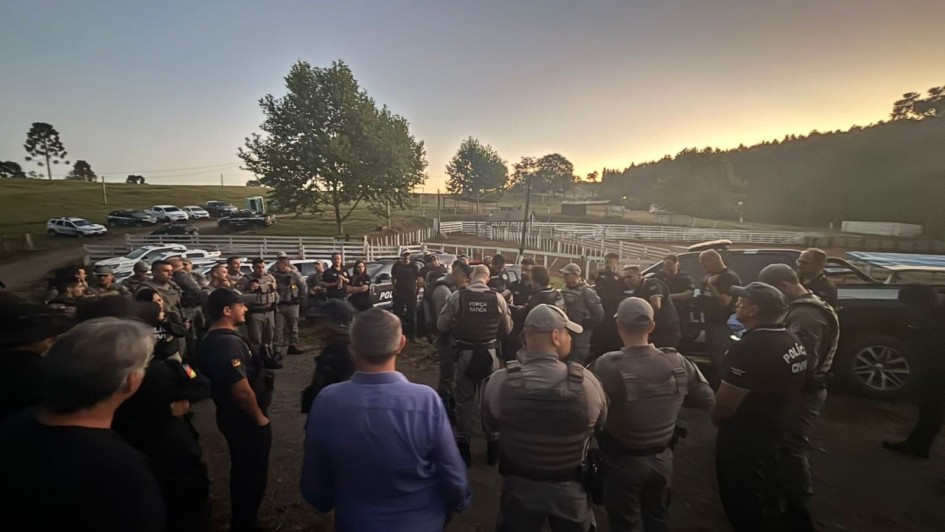 Esta &eacute; uma descri&ccedil;&atilde;o detalhada da imagem para fins de acessibilidade:

Descri&ccedil;&atilde;o Geral
A imagem mostra um grande contingente de for&ccedil;as de seguran&ccedil;a reunido em uma &aacute;rea rural durante o amanhecer ou entardecer. O grupo est&aacute; posicionado em um p&aacute;tio de terra batida, sugerindo o in&iacute;cio ou o encerramento de uma opera&ccedil;&atilde;o policial coordenada.

Elementos em Destaque
O Grupo: Dezenas de agentes da Brigada Militar e da Pol&iacute;cia Civil do Rio Grande do Sul est&atilde;o dispostos em um grande c&iacute;rculo informal. A maioria est&aacute; de costas ou de perfil para a c&acirc;mera.

Vestimentas e Equipamentos:

Agentes da Brigada Militar utilizam o fardamento cinza caracter&iacute;stico e coletes t&aacute;ticos. Um deles, ao centro, possui a inscri&ccedil;&atilde;o "FOR&Ccedil;A T&Aacute;TICA" no colete.

Agentes da Pol&iacute;cia Civil vestem camisetas ou coletes pretos com a inscri&ccedil;&atilde;o "POL&Iacute;CIA CIVIL" em branco nas costas.

Alguns civis e agentes com roupas casuais tamb&eacute;m integram o c&iacute;rculo.

Ve&iacute;culos: Ao fundo, diversas viaturas policiais (caminhonetes e SUVs nas cores branca e preta) est&atilde;o estacionadas de forma organizada pr&oacute;ximas a uma cerca de madeira branca.

Cen&aacute;rio e Ilumina&ccedil;&atilde;o
Ambiente: A cena ocorre em uma zona rural com gramados, &aacute;rvores esparsas e uma estrutura que se assemelha a uma mangueira ou curral ao fundo.

C&eacute;u e Luz: O horizonte apresenta um brilho dourado e alaranjado intenso, t&iacute;pico do sol nascente ou poente, contrastando com o azul suave do restante do c&eacute;u. A ilumina&ccedil;&atilde;o &eacute; baixa, criando silhuetas e uma atmosfera de seriedade e prontid&atilde;o.
