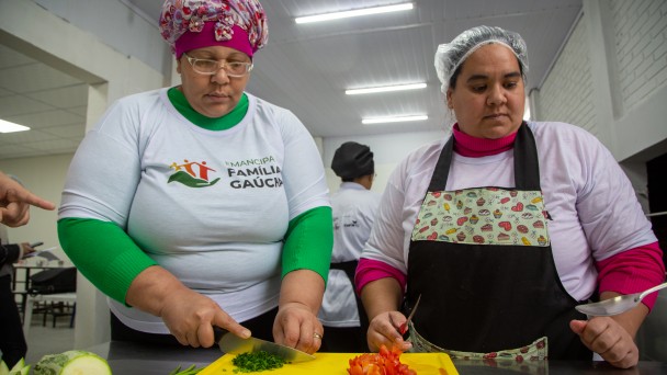 Duas mulheres vestindo aventais e toucas est&atilde;o picando tomates e temperos em uma bancada. Uma veste camiseta identificada com a marca do projeto Emancipa Fam&iacute;lia Ga&uacute;cha. 
