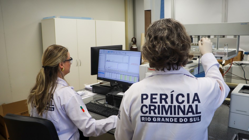 Duas pessoas vestindo jalecos est&atilde;o trabalhando em an&aacute;lises laboratoriais, em frente a um computador. Nas costas do jaleco branco, se l&ecirc; "Per&iacute;cia Criminal Rio Grande do Sul".