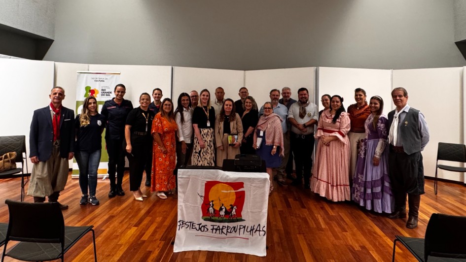 Foto dos instegrantes da Comiss&atilde;o dos Festejos Farroupilhas em meia lua com a bandeira dos Festejos &agrave; frente.