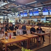 Vis&atilde;o Geral
A imagem captura uma palestra ou apresenta&ccedil;&atilde;o realizada em um ambiente informal e moderno, semelhante a um bar ou restaurante. Uma mulher est&aacute; em p&eacute; &agrave; direita, falando para uma plateia sentada em mesas de madeira.

A Oradora e sua A&ccedil;&atilde;o
Posicionamento: No lado direito da imagem, uma mulher de cabelos castanhos ondulados de comprimento m&eacute;dio est&aacute; de p&eacute; sobre um pequeno tablado ou &aacute;rea de destaque.

Vestimenta: Ela veste um vestido longo, branco e fluido, sem mangas.

A&ccedil;&atilde;o: Ela segura um microfone preto com a m&atilde;o direita e faz um gesto aberto com a m&atilde;o esquerda enquanto fala, demonstrando uma postura comunicativa e engajada.

O P&uacute;blico e o Ambiente
A Plateia: Diversas pessoas de idades variadas est&atilde;o sentadas em mesas de madeira clara, distribu&iacute;das pelo sal&atilde;o. Elas est&atilde;o voltadas para a oradora, com express&otilde;es de aten&ccedil;&atilde;o. Algumas pessoas t&ecirc;m copos, pap&eacute;is e celulares sobre as mesas.

O Local: O estabelecimento possui um design industrial e contempor&acirc;neo. O teto &eacute; alto, com vigas met&aacute;licas escuras, lumin&aacute;rias pendentes em formato de c&uacute;pula e plantas decorativas suspensas em cestos.

O Fundo: Ao fundo, h&aacute; um balc&atilde;o de bar iluminado com prateleiras repletas de garrafas de bebidas. Acima do bar, quatro grandes televisores de tela plana est&atilde;o ligados, exibindo o que parecem ser transmiss&otilde;es de competi&ccedil;&otilde;es esportivas.

Ilumina&ccedil;&atilde;o: A luz do ambiente &eacute; quente e suave, criando uma atmosfera acolhedora para o evento.