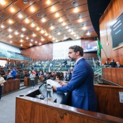 Homem de terno azul fala em um p&uacute;lpito dentro de um grande plen&aacute;rio, com v&aacute;rias pessoas assistindo e tirando fotos.
