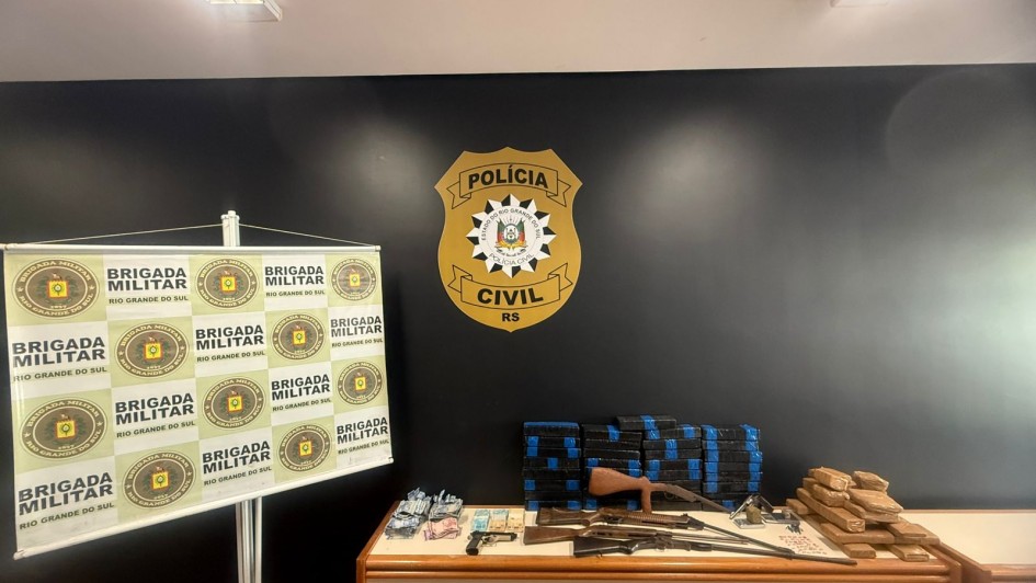 Esta &eacute; uma descri&ccedil;&atilde;o detalhada da imagem para fins de acessibilidade:

Vis&atilde;o Geral
A imagem mostra a exibi&ccedil;&atilde;o de materiais apreendidos em uma opera&ccedil;&atilde;o policial conjunta entre a Pol&iacute;cia Civil e a Brigada Militar. Os itens est&atilde;o organizados sobre uma mesa de madeira contra uma parede preta, em um ambiente interno iluminado por l&acirc;mpadas fluorescentes no teto.

Elementos de Fundo e Identifica&ccedil;&atilde;o
Parede Central: Uma parede pintada de preto fosco exibe, ao centro, um grande emblema dourado e preto da Pol&iacute;cia Civil do RS.

Banner &agrave; Esquerda: Um banner vertical (roll-up) com fundo branco apresenta o bras&atilde;o da Brigada Militar do Rio Grande do Sul repetido em um padr&atilde;o de grade verde e dourado.

Materiais Apreendidos (sobre a mesa)
Da esquerda para a direita, os itens est&atilde;o dispostos da seguinte forma:

Dinheiro: V&aacute;rios ma&ccedil;os de c&eacute;dulas de Real (notas de diversos valores) organizados em pequenos montes.

Armamento: * Uma pistola preta de pequeno porte.

Tr&ecirc;s armas longas (espingardas ou rifles) com acabamento em madeira e metal, dispostas horizontalmente.

Entorpecentes:

Duas grandes pilhas de tijolos retangulares envoltos em fita adesiva azul, totalizando dezenas de unidades.

Uma pilha menor de tijolos retangulares envoltos em fita adesiva parda (cor de papel&atilde;o), localizada no canto direito da mesa.

Contexto
A imagem ilustra os resultados da opera&ccedil;&atilde;o "Ver&atilde;o Sem Drogas", focada no combate ao tr&aacute;fico de entorpecentes na regi&atilde;o do Litoral Norte do estado.