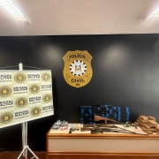 Esta &eacute; uma descri&ccedil;&atilde;o detalhada da imagem para fins de acessibilidade:

Vis&atilde;o Geral
A imagem mostra a exibi&ccedil;&atilde;o de materiais apreendidos em uma opera&ccedil;&atilde;o policial conjunta entre a Pol&iacute;cia Civil e a Brigada Militar. Os itens est&atilde;o organizados sobre uma mesa de madeira contra uma parede preta, em um ambiente interno iluminado por l&acirc;mpadas fluorescentes no teto.

Elementos de Fundo e Identifica&ccedil;&atilde;o
Parede Central: Uma parede pintada de preto fosco exibe, ao centro, um grande emblema dourado e preto da Pol&iacute;cia Civil do RS.

Banner &agrave; Esquerda: Um banner vertical (roll-up) com fundo branco apresenta o bras&atilde;o da Brigada Militar do Rio Grande do Sul repetido em um padr&atilde;o de grade verde e dourado.

Materiais Apreendidos (sobre a mesa)
Da esquerda para a direita, os itens est&atilde;o dispostos da seguinte forma:

Dinheiro: V&aacute;rios ma&ccedil;os de c&eacute;dulas de Real (notas de diversos valores) organizados em pequenos montes.

Armamento: * Uma pistola preta de pequeno porte.

Tr&ecirc;s armas longas (espingardas ou rifles) com acabamento em madeira e metal, dispostas horizontalmente.

Entorpecentes:

Duas grandes pilhas de tijolos retangulares envoltos em fita adesiva azul, totalizando dezenas de unidades.

Uma pilha menor de tijolos retangulares envoltos em fita adesiva parda (cor de papel&atilde;o), localizada no canto direito da mesa.

Contexto
A imagem ilustra os resultados da opera&ccedil;&atilde;o "Ver&atilde;o Sem Drogas", focada no combate ao tr&aacute;fico de entorpecentes na regi&atilde;o do Litoral Norte do estado.