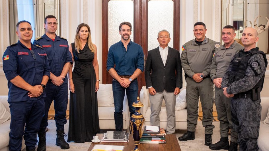 Esta &eacute; uma descri&ccedil;&atilde;o detalhada da imagem para fins de acessibilidade:

Vis&atilde;o Geral
A imagem registra um momento oficial em que oito pessoas &mdash; sete homens e uma mulher &mdash; posam para uma fotografia em um ambiente luxuoso e formal. O grupo est&aacute; organizado em uma linha reta, transmitindo uma atmosfera de coopera&ccedil;&atilde;o institucional.

Pessoas e Vestimentas (da esquerda para a direita)
Bombeiros Militares: Os dois primeiros homens &agrave; esquerda vestem uniformes operacionais azul-marinho com detalhes e identifica&ccedil;&otilde;es em vermelho, t&iacute;picos do Corpo de Bombeiros.

Civil: Uma mulher com cabelos loiros longos veste um vestido preto longo e elegante.

Governador: No centro da imagem, Eduardo Leite veste uma camisa social azul-marinho com as mangas levemente dobradas e cal&ccedil;a jeans escura.

Civil/Autoridade: &Agrave; direita do governador, um homem de cabelos grisalhos veste um blazer preto, camisa branca e cal&ccedil;a cinza-clara.

Brigada Militar: Os dois homens seguintes vestem fardas cinza-oliva da Brigada Militar, com diversos distintivos e ins&iacute;gnias.

T&aacute;tico: Na extremidade direita, um homem veste um uniforme t&aacute;tico camuflado em tons de cinza e preto, equipado com colete operacional.

Cen&aacute;rio e Composi&ccedil;&atilde;o
O Ambiente: O encontro ocorre em uma sala de recep&ccedil;&atilde;o cl&aacute;ssica, possivelmente no Pal&aacute;cio Piratini. O fundo apresenta uma grande porta dupla de madeira com pain&eacute;is de vidro fosco, paredes brancas decoradas com boiseries (molduras de gesso) e espelhos laterais.

Mobili&aacute;rio: Nas laterais, veem-se partes de sof&aacute;s brancos. &Agrave; frente do grupo, h&aacute; uma mesa de centro de madeira escura contendo livros, uma pequena x&iacute;cara de caf&eacute; sobre um pires branco e um vaso decorativo alongado em azul-marinho com detalhes dourados.

Ilumina&ccedil;&atilde;o: A luz &eacute; suave e bem distribu&iacute;da, real&ccedil;ando os detalhes dos uniformes e do ambiente.