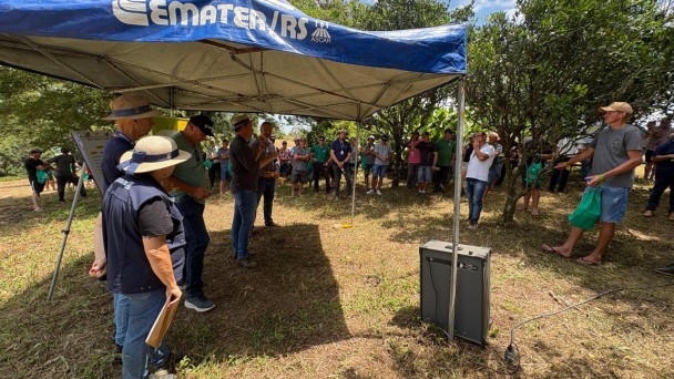 Foto mostra t&eacute;cnicos em baixo de uma tenda da Emater falando para produtores e autoridades que est&atilde;o embaixo da sombra de &aacute;rvores no terreno da propriedade. 