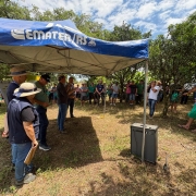 Foto mostra t&eacute;cnicos em baixo de uma tenda da Emater falando para produtores e autoridades que est&atilde;o embaixo da sombra de &aacute;rvores no terreno da propriedade. 