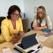 Esta &eacute; uma descri&ccedil;&atilde;o detalhada da imagem para fins de acessibilidade:

Vis&atilde;o Geral
A imagem mostra duas mulheres sentadas &agrave; mesa de um escrit&oacute;rio, em frente a um tablet, durante o que parece ser uma videoconfer&ecirc;ncia ou reuni&atilde;o virtual. A cena transmite uma atmosfera de trabalho focado e coordena&ccedil;&atilde;o institucional.

As Pessoas e suas A&ccedil;&otilde;es
Mulher &agrave; esquerda: Est&aacute; sentada em uma cadeira de escrit&oacute;rio preta, vestindo um blazer amarelo vibrante sobre uma blusa do mesmo tom. Ela usa &oacute;culos de arma&ccedil;&atilde;o vermelha e observa atentamente a tela do tablet. Em seu pulso esquerdo, v&ecirc;-se um rel&oacute;gio inteligente com pulseira clara.

Mulher &agrave; direita: Est&aacute; sentada ao lado, vestindo uma camisa de tecido leve na cor bege. Ela usa &oacute;culos de arma&ccedil;&atilde;o clara e est&aacute; com os bra&ccedil;os cruzados sobre a mesa, mantendo o olhar fixo na tela.

Cen&aacute;rio e Objetos
A Mesa: Sobre a mesa de cor clara, h&aacute; uma pilha de pap&eacute;is e cadernos, com o tablet posicionado horizontalmente em cima deles, servindo como tela principal para a reuni&atilde;o. No canto direito da mesa, h&aacute; uma garrafa t&eacute;rmica ou recipiente alto na cor rosa claro.

Ambiente: O fundo &eacute; composto por uma parede branca e lisa. Ao fundo, &agrave; esquerda, sobre um balc&atilde;o de madeira, percebem-se outros objetos de escrit&oacute;rio e recipientes de &aacute;gua.

Contexto
A imagem ilustra a mobiliza&ccedil;&atilde;o do governo estadual junto aos munic&iacute;pios para o fortalecimento da rede de prote&ccedil;&atilde;o &agrave;s mulheres durante o per&iacute;odo do Carnaval.