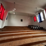 auditório com grandes janelas e cortinas, ao fundo algumas cadeiras dispostas