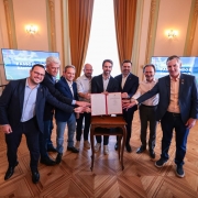 A imagem registra um grupo de pessoas alinhado em frente a uma pequena mesa de madeira posicionada no centro do sal&atilde;o, exibindo um documento aberto com capa vermelha. Todas estendem as m&atilde;os em dire&ccedil;&atilde;o ao documento assinado. 