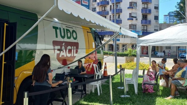 Esta &eacute; uma descri&ccedil;&atilde;o detalhada da imagem para fins de acessibilidade:

Vis&atilde;o Geral
A imagem registra a opera&ccedil;&atilde;o de uma unidade m&oacute;vel do servi&ccedil;o Tudo F&aacute;cil em um ambiente externo urbano, sob luz solar intensa, indicando um dia claro.

O Ve&iacute;culo e o Atendimento
O Ve&iacute;culo: &Agrave; esquerda, um &ocirc;nibus ou van de grande porte est&aacute; estacionado. A pintura do ve&iacute;culo utiliza as cores verde, amarelo e vermelho, com o logotipo "TUDO F&Aacute;CIL" em destaque na parte central branca.

Estrutura de Sombra: Um grande toldo branco est&aacute; estendido da lateral do ve&iacute;culo, cobrindo a &aacute;rea de trabalho.

Equipe: Duas mulheres vestindo camisetas pretas (uniforme) manuseiam notebooks sobre mesas pretas posicionadas junto ao ve&iacute;culo. Uma delas est&aacute; em p&eacute; e a outra sentada.

Organiza&ccedil;&atilde;o: Barreiras de conten&ccedil;&atilde;o com fitas pretas delimitam o espa&ccedil;o de atendimento e organizam a circula&ccedil;&atilde;o.

&Aacute;rea de Espera e P&uacute;blico
Tenda de Espera: &Agrave; direita, h&aacute; uma tenda branca montada sobre um gramado.

Pessoas: Sob a tenda, cerca de cinco pessoas (incluindo adultos e uma crian&ccedil;a) aguardam sentadas em cadeiras de pl&aacute;stico brancas e pretas. Elas vestem roupas leves e casuais.

Cen&aacute;rio de Fundo
Arquitetura: Ao fundo, elevam-se edif&iacute;cios residenciais de v&aacute;rios andares, com sacadas arredondadas e detalhes em azul.

Entorno: H&aacute; &aacute;rvores, vegeta&ccedil;&atilde;o baixa e uma rua pavimentada que contorna o local do evento.