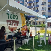 Esta &eacute; uma descri&ccedil;&atilde;o detalhada da imagem para fins de acessibilidade:

Vis&atilde;o Geral
A imagem registra a opera&ccedil;&atilde;o de uma unidade m&oacute;vel do servi&ccedil;o Tudo F&aacute;cil em um ambiente externo urbano, sob luz solar intensa, indicando um dia claro.

O Ve&iacute;culo e o Atendimento
O Ve&iacute;culo: &Agrave; esquerda, um &ocirc;nibus ou van de grande porte est&aacute; estacionado. A pintura do ve&iacute;culo utiliza as cores verde, amarelo e vermelho, com o logotipo "TUDO F&Aacute;CIL" em destaque na parte central branca.

Estrutura de Sombra: Um grande toldo branco est&aacute; estendido da lateral do ve&iacute;culo, cobrindo a &aacute;rea de trabalho.

Equipe: Duas mulheres vestindo camisetas pretas (uniforme) manuseiam notebooks sobre mesas pretas posicionadas junto ao ve&iacute;culo. Uma delas est&aacute; em p&eacute; e a outra sentada.

Organiza&ccedil;&atilde;o: Barreiras de conten&ccedil;&atilde;o com fitas pretas delimitam o espa&ccedil;o de atendimento e organizam a circula&ccedil;&atilde;o.

&Aacute;rea de Espera e P&uacute;blico
Tenda de Espera: &Agrave; direita, h&aacute; uma tenda branca montada sobre um gramado.

Pessoas: Sob a tenda, cerca de cinco pessoas (incluindo adultos e uma crian&ccedil;a) aguardam sentadas em cadeiras de pl&aacute;stico brancas e pretas. Elas vestem roupas leves e casuais.

Cen&aacute;rio de Fundo
Arquitetura: Ao fundo, elevam-se edif&iacute;cios residenciais de v&aacute;rios andares, com sacadas arredondadas e detalhes em azul.

Entorno: H&aacute; &aacute;rvores, vegeta&ccedil;&atilde;o baixa e uma rua pavimentada que contorna o local do evento.