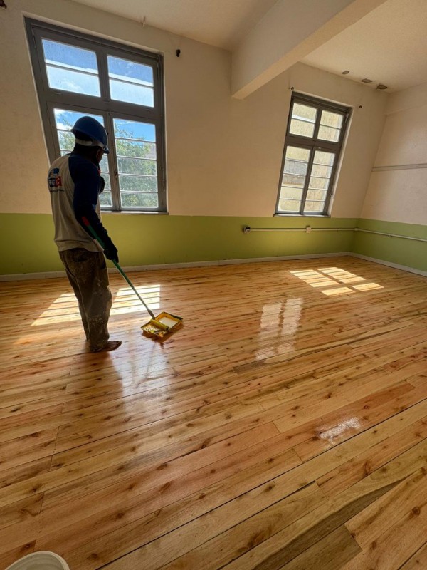 Homem pinta piso de sala com duas grandes janelas. Piso está muito brilhoso. 