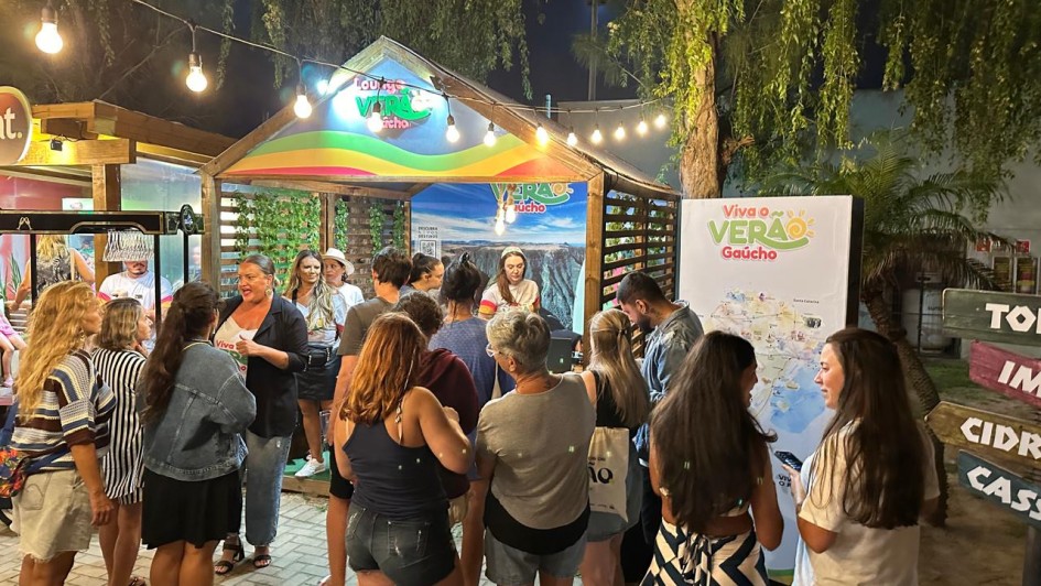A imagem captura uma cena noturna vibrante em um espa&ccedil;o de atendimento ao turista. Diversas pessoas est&atilde;o reunidas em torno de um quiosque de madeira decorado, sob uma ilumina&ccedil;&atilde;o quente e festiva.

O Quiosque e a Ambienta&ccedil;&atilde;o
Estrutura: No centro, h&aacute; um quiosque de madeira com telhado em ponta, identificado como "Lounge Viva o Ver&atilde;o Ga&uacute;cho". O front&atilde;o &eacute; decorado com faixas coloridas (arco-&iacute;ris) e o logotipo da campanha.

Fundo do Quiosque: Um grande painel exibe a foto de uma paisagem natural exuberante, possivelmente os c&acirc;nions da regi&atilde;o, servindo de cen&aacute;rio para o atendimento.

Ilumina&ccedil;&atilde;o: O ambiente &eacute; iluminado por um varal de luzes (l&acirc;mpadas de filamento) que atravessa a parte superior, criando uma atmosfera acolhedora e convidativa.

Sinaliza&ccedil;&atilde;o: &Agrave; direita, h&aacute; um totem branco com o t&iacute;tulo "Viva o Ver&atilde;o Ga&uacute;cho" e um mapa detalhado da regi&atilde;o. Mais &agrave; direita, um poste de madeira indica dire&ccedil;&otilde;es para locais como "CIDREIRA" e "CASSINO".

As Pessoas
Intera&ccedil;&atilde;o: Um grupo diverso de cerca de 10 a 12 pessoas, vestindo roupas leves de ver&atilde;o (camisetas, bermudas e vestidos), circula pelo espa&ccedil;o.

Atendimento: Algumas pessoas est&atilde;o voltadas para o interior do quiosque conversando com uma atendente, enquanto outras observam o mapa ou conversam entre si em pequenos grupos.

Atitude: O clima parece descontra&iacute;do e social, t&iacute;pico de um evento de lazer ou ponto de informa&ccedil;&otilde;es tur&iacute;sticas.

Detalhes Adicionais
Piso: O ch&atilde;o &eacute; revestido com blocos de concreto intertravados.

Vegeta&ccedil;&atilde;o: &Aacute;rvores com folhagens finas e uma palmeira ao fundo emolduram a cena contra o c&eacute;u escuro da noite.