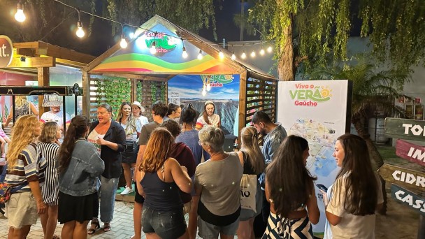 A imagem captura uma cena noturna vibrante em um espa&ccedil;o de atendimento ao turista. Diversas pessoas est&atilde;o reunidas em torno de um quiosque de madeira decorado, sob uma ilumina&ccedil;&atilde;o quente e festiva.

O Quiosque e a Ambienta&ccedil;&atilde;o
Estrutura: No centro, h&aacute; um quiosque de madeira com telhado em ponta, identificado como "Lounge Viva o Ver&atilde;o Ga&uacute;cho". O front&atilde;o &eacute; decorado com faixas coloridas (arco-&iacute;ris) e o logotipo da campanha.

Fundo do Quiosque: Um grande painel exibe a foto de uma paisagem natural exuberante, possivelmente os c&acirc;nions da regi&atilde;o, servindo de cen&aacute;rio para o atendimento.

Ilumina&ccedil;&atilde;o: O ambiente &eacute; iluminado por um varal de luzes (l&acirc;mpadas de filamento) que atravessa a parte superior, criando uma atmosfera acolhedora e convidativa.

Sinaliza&ccedil;&atilde;o: &Agrave; direita, h&aacute; um totem branco com o t&iacute;tulo "Viva o Ver&atilde;o Ga&uacute;cho" e um mapa detalhado da regi&atilde;o. Mais &agrave; direita, um poste de madeira indica dire&ccedil;&otilde;es para locais como "CIDREIRA" e "CASSINO".

As Pessoas
Intera&ccedil;&atilde;o: Um grupo diverso de cerca de 10 a 12 pessoas, vestindo roupas leves de ver&atilde;o (camisetas, bermudas e vestidos), circula pelo espa&ccedil;o.

Atendimento: Algumas pessoas est&atilde;o voltadas para o interior do quiosque conversando com uma atendente, enquanto outras observam o mapa ou conversam entre si em pequenos grupos.

Atitude: O clima parece descontra&iacute;do e social, t&iacute;pico de um evento de lazer ou ponto de informa&ccedil;&otilde;es tur&iacute;sticas.

Detalhes Adicionais
Piso: O ch&atilde;o &eacute; revestido com blocos de concreto intertravados.

Vegeta&ccedil;&atilde;o: &Aacute;rvores com folhagens finas e uma palmeira ao fundo emolduram a cena contra o c&eacute;u escuro da noite.