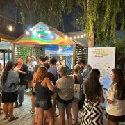 A imagem captura uma cena noturna vibrante em um espa&ccedil;o de atendimento ao turista. Diversas pessoas est&atilde;o reunidas em torno de um quiosque de madeira decorado, sob uma ilumina&ccedil;&atilde;o quente e festiva.

O Quiosque e a Ambienta&ccedil;&atilde;o
Estrutura: No centro, h&aacute; um quiosque de madeira com telhado em ponta, identificado como "Lounge Viva o Ver&atilde;o Ga&uacute;cho". O front&atilde;o &eacute; decorado com faixas coloridas (arco-&iacute;ris) e o logotipo da campanha.

Fundo do Quiosque: Um grande painel exibe a foto de uma paisagem natural exuberante, possivelmente os c&acirc;nions da regi&atilde;o, servindo de cen&aacute;rio para o atendimento.

Ilumina&ccedil;&atilde;o: O ambiente &eacute; iluminado por um varal de luzes (l&acirc;mpadas de filamento) que atravessa a parte superior, criando uma atmosfera acolhedora e convidativa.

Sinaliza&ccedil;&atilde;o: &Agrave; direita, h&aacute; um totem branco com o t&iacute;tulo "Viva o Ver&atilde;o Ga&uacute;cho" e um mapa detalhado da regi&atilde;o. Mais &agrave; direita, um poste de madeira indica dire&ccedil;&otilde;es para locais como "CIDREIRA" e "CASSINO".

As Pessoas
Intera&ccedil;&atilde;o: Um grupo diverso de cerca de 10 a 12 pessoas, vestindo roupas leves de ver&atilde;o (camisetas, bermudas e vestidos), circula pelo espa&ccedil;o.

Atendimento: Algumas pessoas est&atilde;o voltadas para o interior do quiosque conversando com uma atendente, enquanto outras observam o mapa ou conversam entre si em pequenos grupos.

Atitude: O clima parece descontra&iacute;do e social, t&iacute;pico de um evento de lazer ou ponto de informa&ccedil;&otilde;es tur&iacute;sticas.

Detalhes Adicionais
Piso: O ch&atilde;o &eacute; revestido com blocos de concreto intertravados.

Vegeta&ccedil;&atilde;o: &Aacute;rvores com folhagens finas e uma palmeira ao fundo emolduram a cena contra o c&eacute;u escuro da noite.