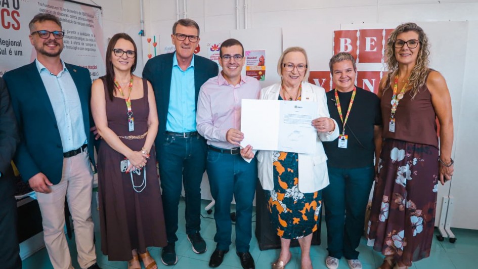 Esta &eacute; uma descri&ccedil;&atilde;o detalhada da imagem para fins de acessibilidade:

Vis&atilde;o Geral
A imagem mostra um grupo de sete pessoas (quatro mulheres e tr&ecirc;s homens) posando para uma foto oficial em um ambiente interno. Eles est&atilde;o alinhados lado a lado, e o registro captura um momento de celebra&ccedil;&atilde;o institucional.

Pessoas e A&ccedil;&otilde;es
Destaque Central: No centro do grupo, uma mulher de cabelos loiros curtos e &oacute;culos, vestindo um blazer branco e uma saia com estampa floral colorida, segura um documento oficial assinado. Ao lado dela, um homem de camisa rosa clara e cal&ccedil;a azul tamb&eacute;m segura a outra extremidade do papel, ambos sorrindo para a c&acirc;mera.

Composi&ccedil;&atilde;o do Grupo:

&Agrave; esquerda do centro, h&aacute; um homem alto de blazer azul marinho e camisa azul clara, seguido por uma mulher de vestido marrom e &oacute;culos, e na extremidade esquerda, um homem jovem de &oacute;culos, blazer escuro e cal&ccedil;a bege.

&Agrave; direita do centro, est&atilde;o uma mulher de camiseta preta e &oacute;culos, e na extremidade direita, uma mulher de blusa marrom sem mangas e saia longa com estampa floral escura.

Identifica&ccedil;&atilde;o: Quase todos os presentes utilizam crach&aacute;s pendurados no pesco&ccedil;o com cord&otilde;es coloridos, indicando serem representantes oficiais ou t&eacute;cnicos.

O Ambiente
Fundo: Atr&aacute;s do grupo, h&aacute; pain&eacute;is brancos de divis&oacute;ria. No lado esquerdo, um banner exibe o logotipo do HEMOCS (Hemocentro Regional de Caxias do Sul) e textos sobre a import&acirc;ncia da doa&ccedil;&atilde;o de sangue.

Detalhes: O ch&atilde;o &eacute; de cer&acirc;mica em tons de verde claro e a ilumina&ccedil;&atilde;o &eacute; uniforme, t&iacute;pica de um ambiente de escrit&oacute;rio ou sala de reuni&otilde;es oficial.