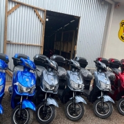 Esta &eacute; uma descri&ccedil;&atilde;o detalhada da imagem para fins de acessibilidade:

Vis&atilde;o Geral
A imagem apresenta uma fileira de motocicletas el&eacute;tricas ou scooters novas, estacionadas em frente a uma instala&ccedil;&atilde;o da Pol&iacute;cia Penal do Rio Grande do Sul.

As Motocicletas
Cores e Modelos: Est&atilde;o alinhadas cerca de oito motocicletas em tr&ecirc;s cores distintas: azul vibrante &agrave; esquerda, cinza met&aacute;lico ao centro e vermelho &agrave; direita.

Detalhes: Todas possuem um ba&uacute; de carga preto (top case) instalado na parte traseira. Algumas ainda mant&ecirc;m o pl&aacute;stico de prote&ccedil;&atilde;o nos assentos, indicando serem ve&iacute;culos novos.

O Ambiente
Edifica&ccedil;&atilde;o: As motos est&atilde;o estacionadas em um p&aacute;tio de brita em frente a um galp&atilde;o com paredes de metal galvanizado e uma porta de correr parcialmente aberta.

Identifica&ccedil;&atilde;o Oficial: Na parede lateral clara, &agrave; direita, destaca-se o bras&atilde;o oficial amarelo e verde da Pol&iacute;cia Penal RS.

Contexto: A cena sugere a moderniza&ccedil;&atilde;o da frota ou a implementa&ccedil;&atilde;o de novos recursos de mobilidade para o sistema prisional, em conformidade com as normas mencionadas no t&iacute;tulo do arquivo.