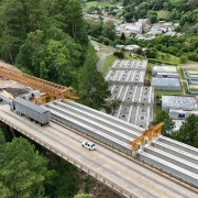Esta é uma descrição detalhada da imagem para fins de acessibilidade:

Visão Geral
A imagem apresenta uma vista aérea de grande ângulo (provavelmente capturada por um drone) de uma importante obra de infraestrutura rodoviária. O cenário mostra a construção de novos pilares para uma ponte em meio a um vale de vegetação densa e preservada.

A Ponte e a Obra
Ponte Existente: Uma longa ponte de concreto cinza atravessa o vale diagonalmente, da esquerda para a direita. Veículos leves podem ser vistos circulando sobre ela.

Novas Estruturas: Paralelamente à ponte atual, estão sendo erguidos novos pilares de sustentação em concreto.

Atividade de Construção:

No topo dos novos pilares, há plataformas de trabalho cercadas por andaimes e formas de madeira. É possível avistar operários em uniformes laranjas trabalhando nessas alturas.

Na base da estrutura central, está posicionado um caminhão bomba de concreto branco com braços estendidos.

Estruturas metálicas amarelas (provavelmente treliças de lançamento ou guindastes) estão em uso para o posicionamento de vigas na extremidade inferior direita da obra.

Preparação do Terreno: Áreas de encosta próximas à cabeceira da ponte exibem terraplanagem e contenções feitas com telas de proteção laranjas.

O Entorno Natural
Vegetação: O vale é coberto por uma floresta verde exuberante e contínua, que contrasta com o tom cinza das estruturas de concreto.

Geografia: O terreno é acidentado, com morros íngremes. Um caminho de terra serpenteia o fundo do vale, próximo ao leito do arroio.