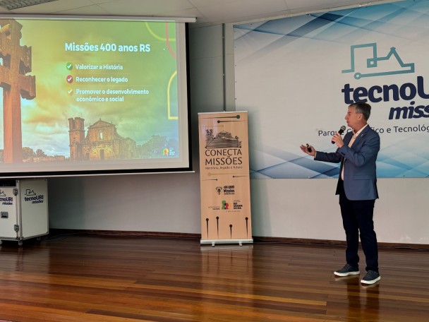 Secretário Eduardo Loureiro, homem branco, vestindo terno azul claro e calça preta está de pé em um palco e gesticula com a mão esquerda em direção a tela e segura microfone com a mão direita. Na tela, aparece uma imagem estilizada das ruínas de São Miguel das Missões
