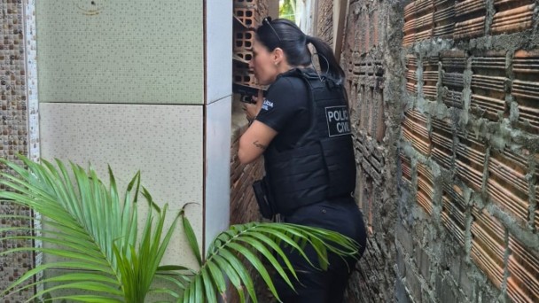 Esta &eacute; uma descri&ccedil;&atilde;o detalhada da imagem para fins de acessibilidade:

Vis&atilde;o Geral
A imagem mostra uma policial civil em a&ccedil;&atilde;o t&aacute;tica durante a Opera&ccedil;&atilde;o Carnaval Seguro. A cena ocorre em um ambiente urbano estreito, capturando um momento de progress&atilde;o policial entre edifica&ccedil;&otilde;es.

A Policial e a A&ccedil;&atilde;o
Identifica&ccedil;&atilde;o e Traje: A policial, uma mulher de pele clara e cabelos escuros presos em um rabo de cavalo, veste o uniforme da Pol&iacute;cia Civil. Ela utiliza uma camiseta preta sob um colete t&aacute;tico bal&iacute;stico tamb&eacute;m preto, onde se l&ecirc; a inscri&ccedil;&atilde;o da institui&ccedil;&atilde;o.

Postura e Equipamento: Posicionada de perfil, ela segura uma pistola preta em punho, mantendo uma postura de alerta e foco enquanto observa atrav&eacute;s de uma abertura ou fresta na parede.

Detalhe Particular: No antebra&ccedil;o esquerdo da policial, &eacute; poss&iacute;vel notar uma pequena tatuagem com a palavra "Love".

O Cen&aacute;rio
Arquitetura: A a&ccedil;&atilde;o se desenvolve em um corredor estreito. De um lado, h&aacute; uma parede de tijolos cer&acirc;micos aparentes e sem reboco; do outro, uma parede com revestimento de azulejos claros e quadrados.

Vegeta&ccedil;&atilde;o: No canto inferior esquerdo da imagem, as folhas verdes de uma pequena planta (tipo palmeira) aparecem em primeiro plano, adicionando um elemento org&acirc;nico &agrave; cena urbana.