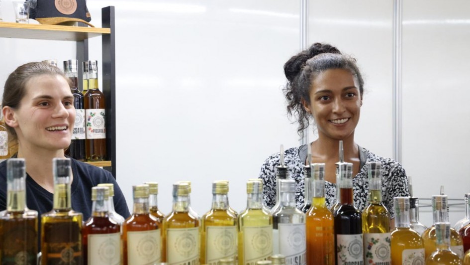 Foto de duas mulheres atr&aacute;s de uma banca de bebidas. Elas sorriem.
