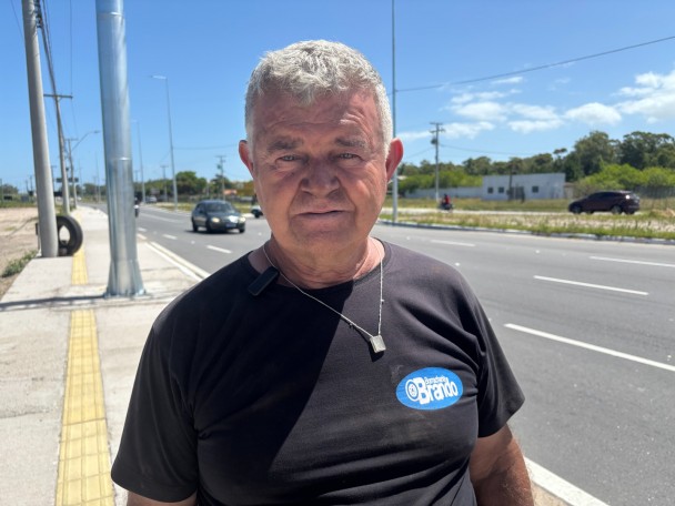 Foto em ambiente aberto, na calçada de uma estrada, de um homem idoso, em primeiro plano. Ele tem cabelos brancos, usa camiseta preta e olha para a câmera.