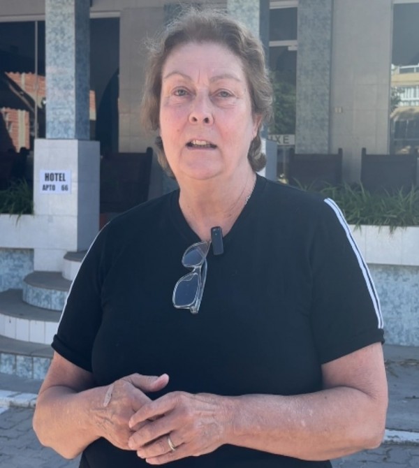 Foto em plano americano, ao ar livre, de uma mulher idosa, de cabelos castanhos. Ela usa camiseta preta e olha para a câmera.