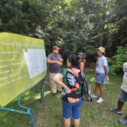 Foto em &aacute;rea vegetal de pessoal de p&eacute; ouvindo um guia, que est&aacute; ao lado de uma placa.